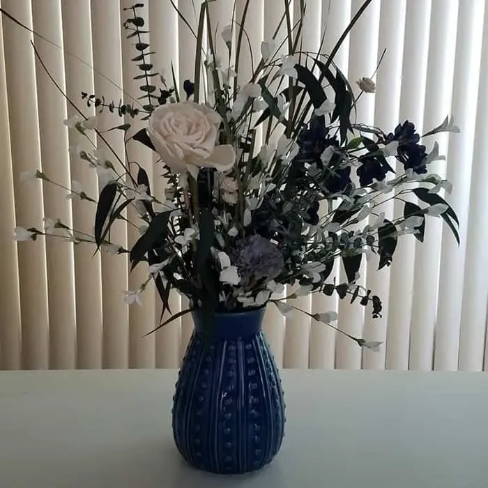 Blue vase with white, blue, and green flowers in front of a window with vertical blinds.