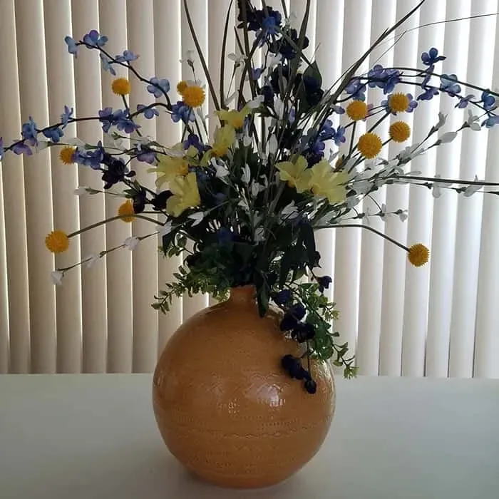 Round, orange vase with a floral arrangement of yellow, blue, and white flowers on a white table.