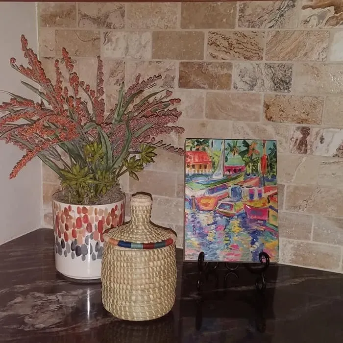 A floral arrangement, wicker jar, and painting on a stand on a dark countertop with a brick tile backsplash.