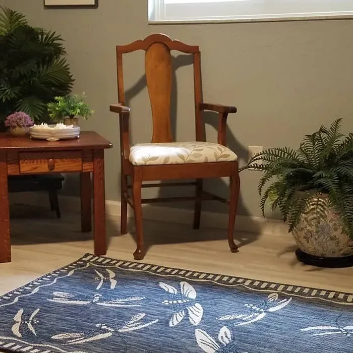 Wooden chair with patterned seat, side table, blue rug, and plants in a room with a gray wall.