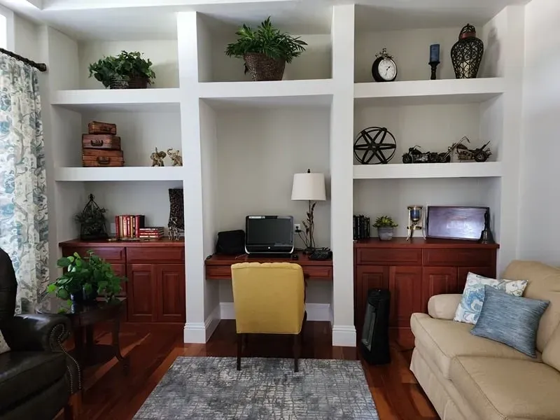Built-in shelving with desk space, containing plants, décor, and a laptop. Yellow chair on a rug, hardwood floors.