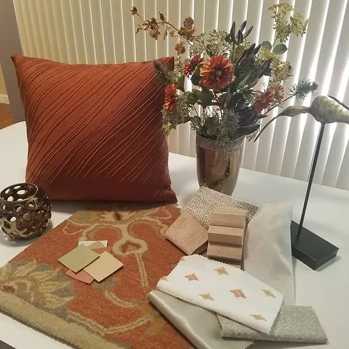 Orange pillow, rug, flowers, and fabric swatches on a white table, showcasing interior design elements.
