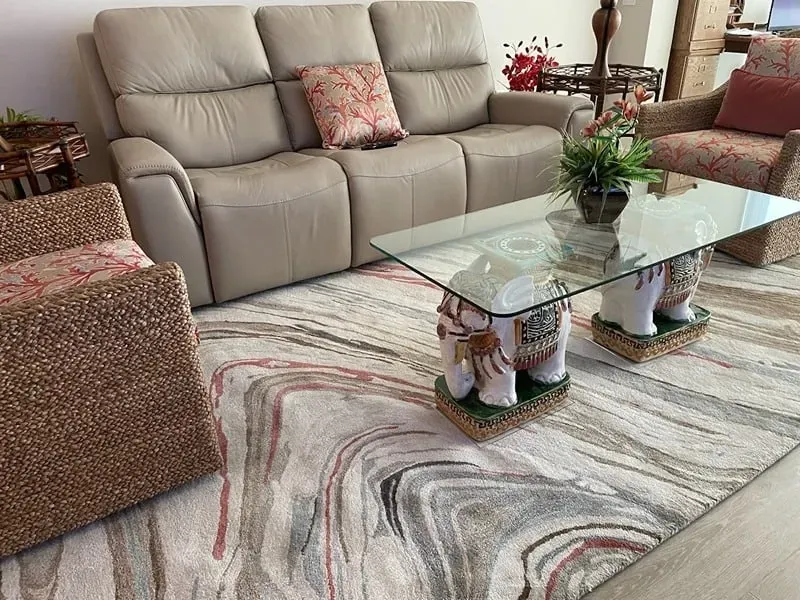 Living room with beige sofa, patterned rug, glass table on elephant legs, and wicker chairs.
