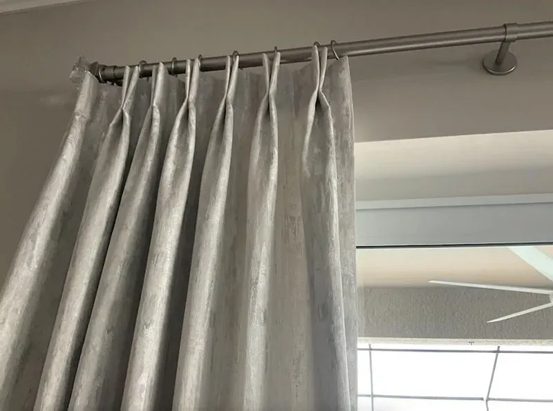 Silver pleated curtains on a metal rod, in front of a window and a ceiling fan.