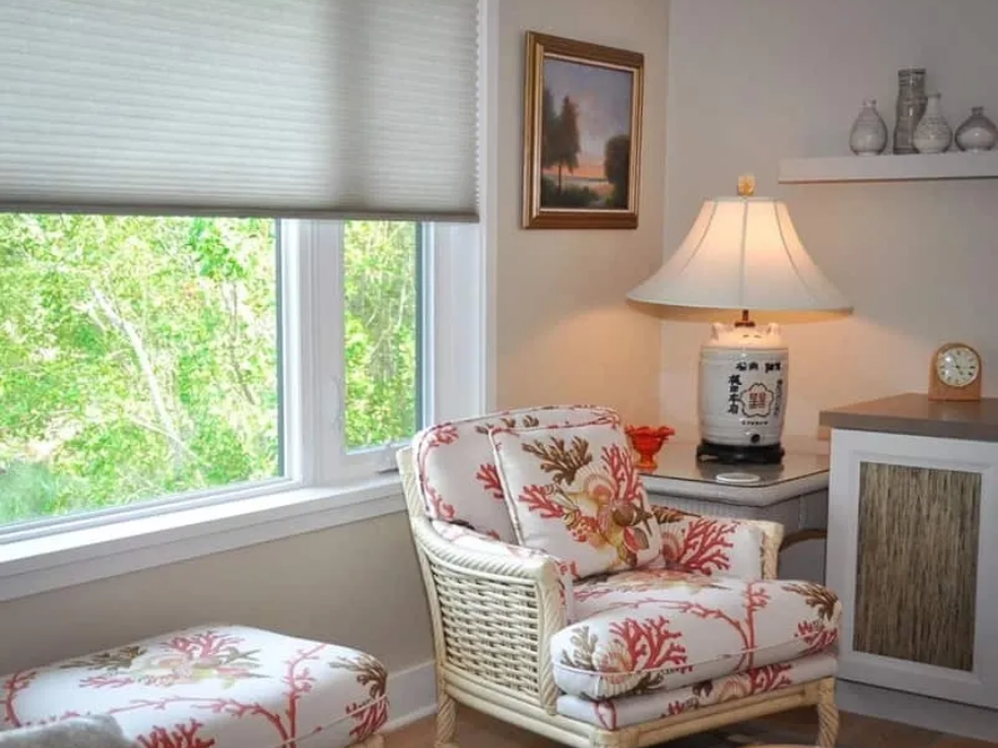 Cozy corner with patterned armchair, ottoman, lamp, and window overlooking green foliage.