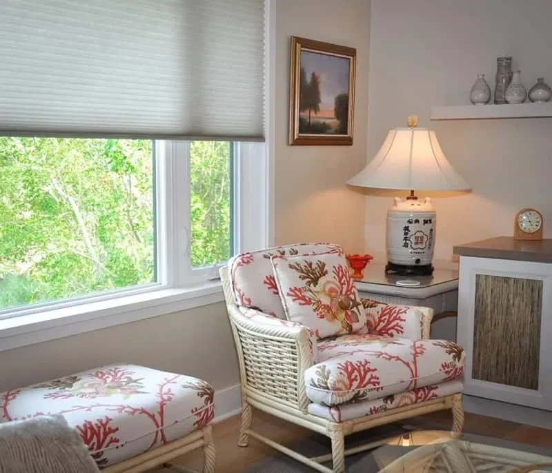 Cozy room with armchair, ottoman, and window overlooking trees. Features floral fabric and a lit lamp.
