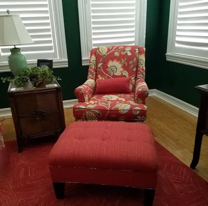Cozy room with floral armchair, ottoman, lamp, and side table on a red rug. Green walls and shuttered windows.