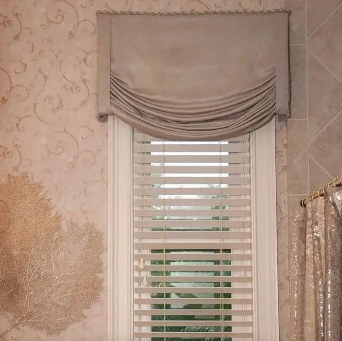 Window with beige draped valance above horizontal blinds, set in a room with patterned wallpaper.