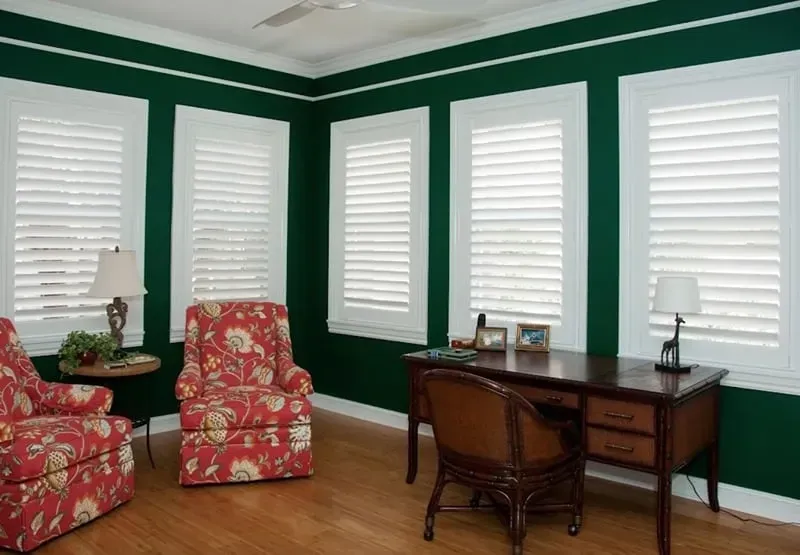 A room with white shuttered windows, dark green walls, and floral armchairs. A desk and lamp are also present.