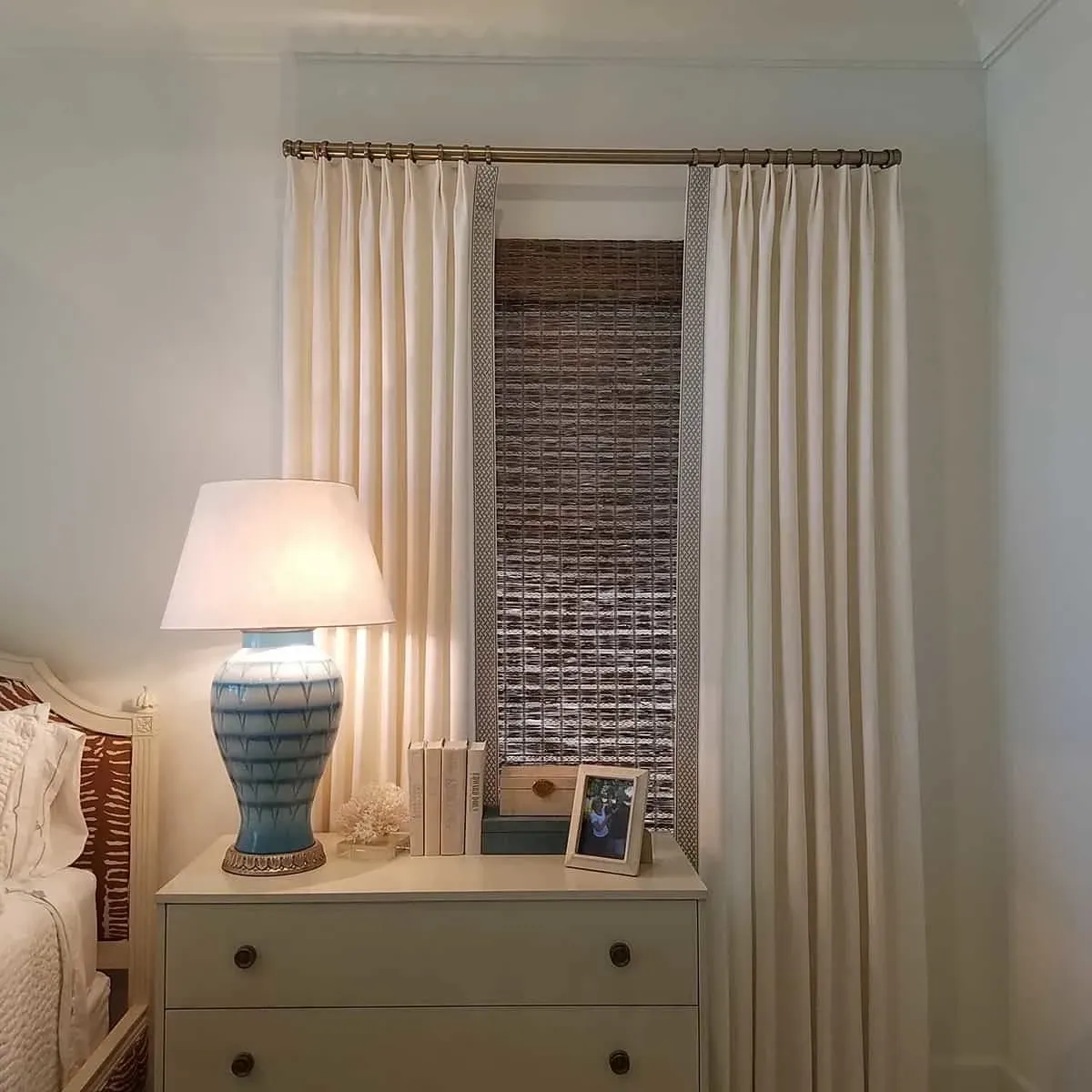 Bedroom window with light cream curtains, bamboo blinds, and a blue lamp on a white dresser.