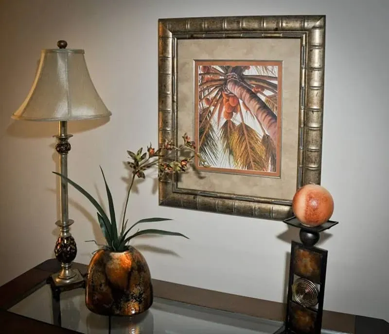 Table with lamp, plant in decorative vase, framed artwork of palm tree, and candle holder.