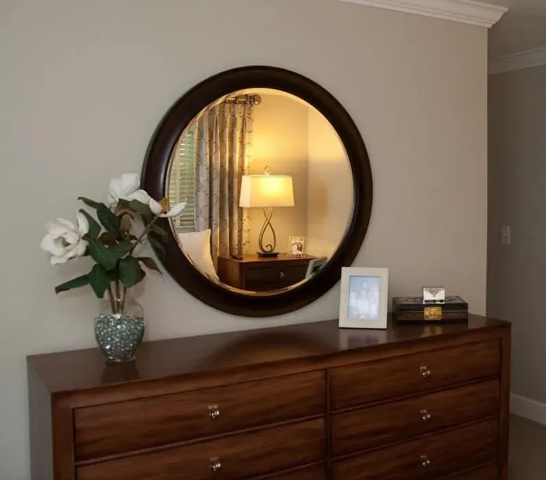 Wooden dresser with round mirror reflecting a lamp and window. Flowers and picture frame on top.