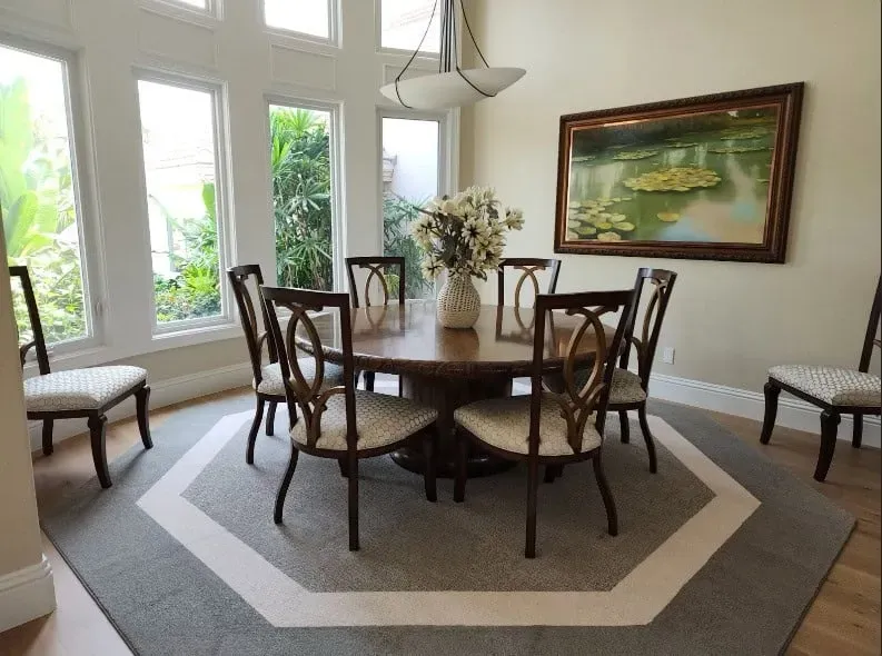 Dining room with round table, eight chairs, large windows, and floral painting.