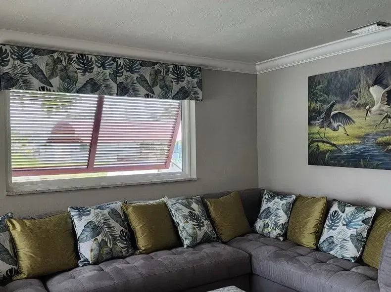 Living room with a gray sectional sofa, floral pillows, and a window with blinds.