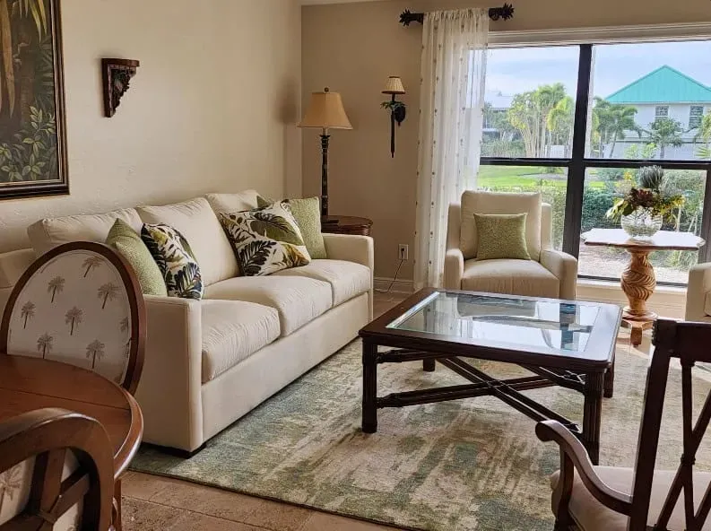 Living room with off-white sofa, armchairs, coffee table, and area rug. Large window overlooks a green landscape.
