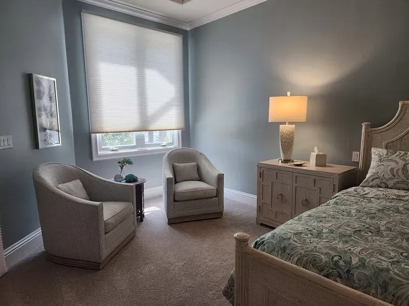 Cozy bedroom with blue walls, two armchairs, a bed, a window with shades, and a lamp on a nightstand.