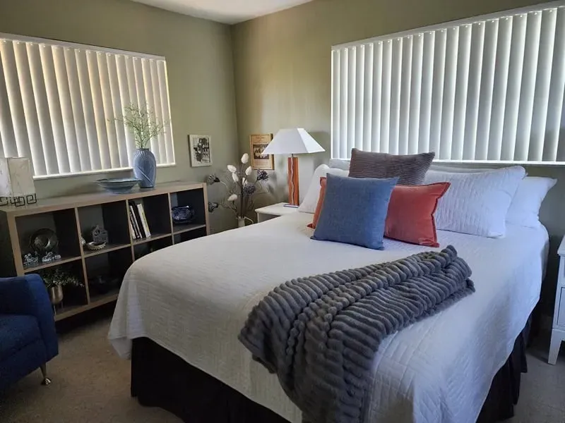 Bedroom with white bed, gray throw, and assorted pillows, beside a table lamp.