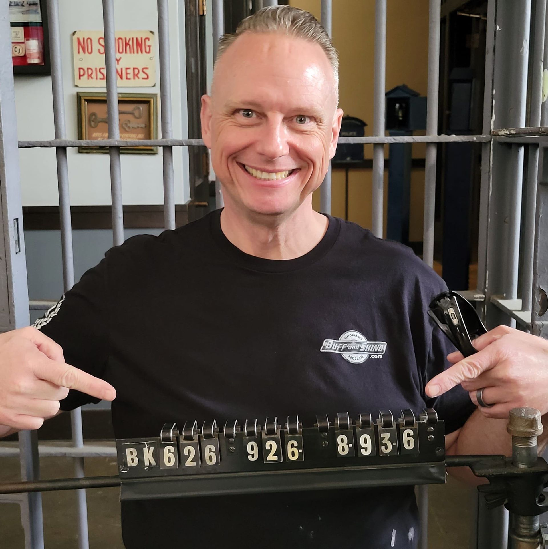 Man smiling, pointing at jail cell lock numbers: BK626, 926, 8936. Jail setting, black t-shirt.