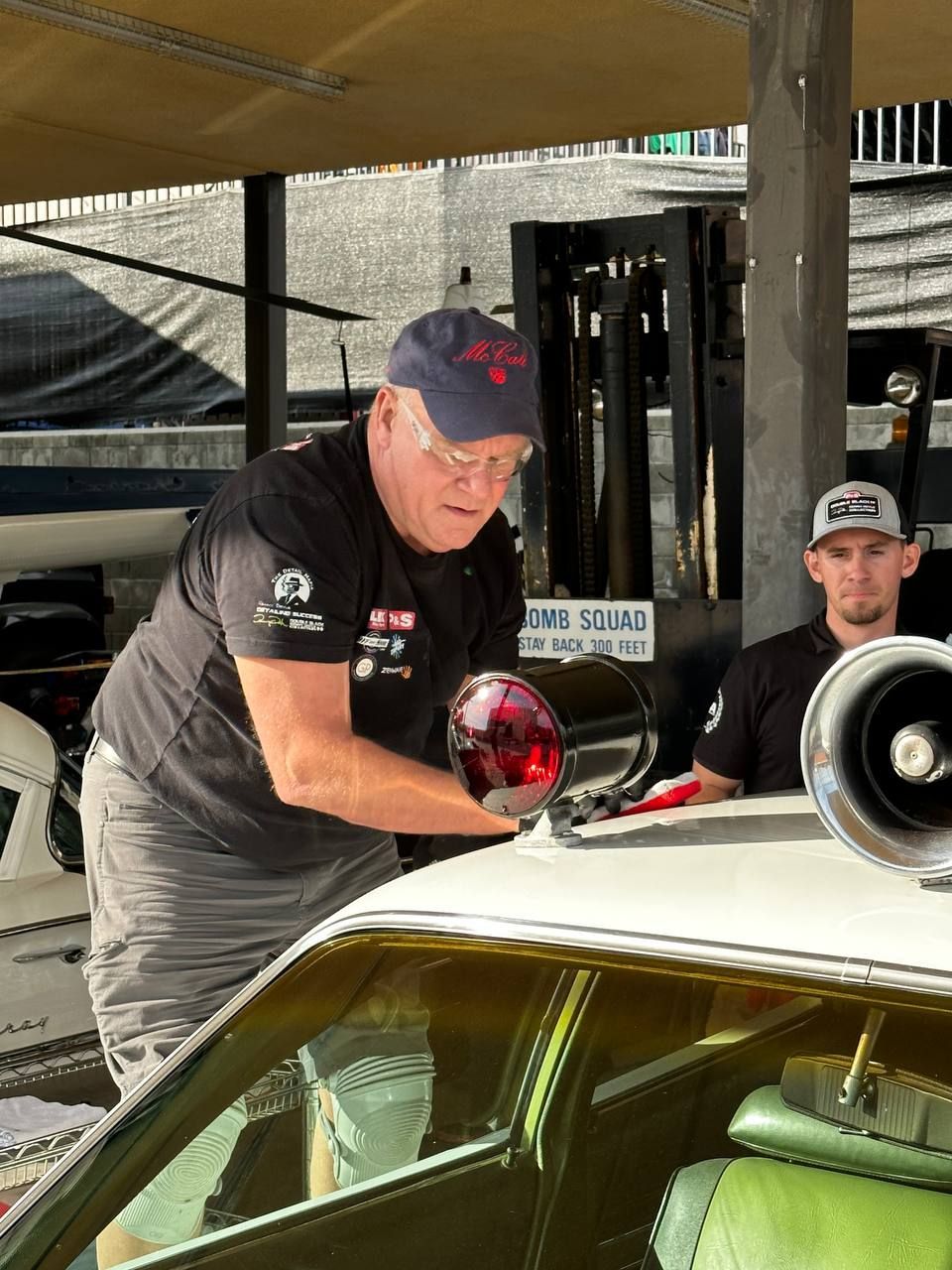 Man in black shirt installs red light on white car. Another man sits behind him.