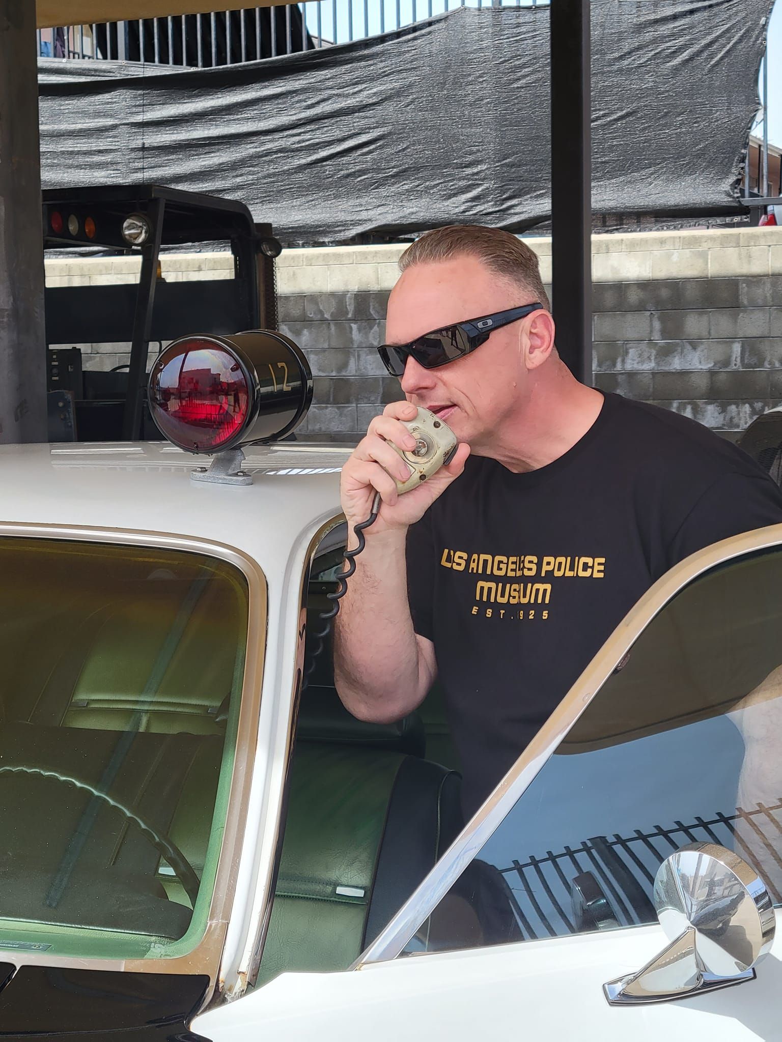 Man in sunglasses on a police car, talking into a radio. Outdoors, bright sunlight.