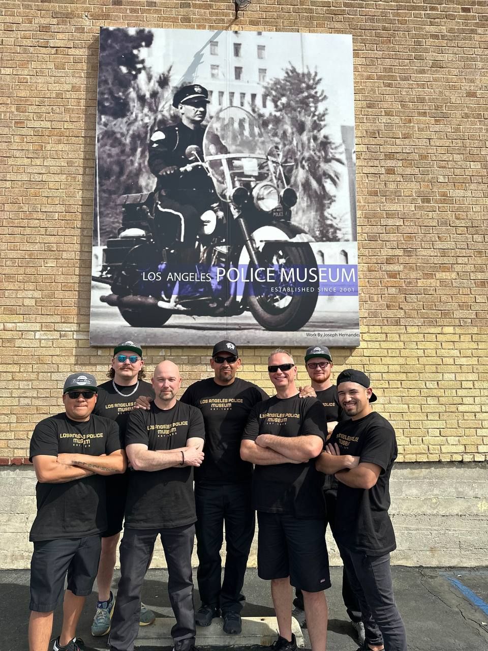 Group of men pose in front of the Los Angeles Police Museum sign, a photo of a cop on a motorcycle.