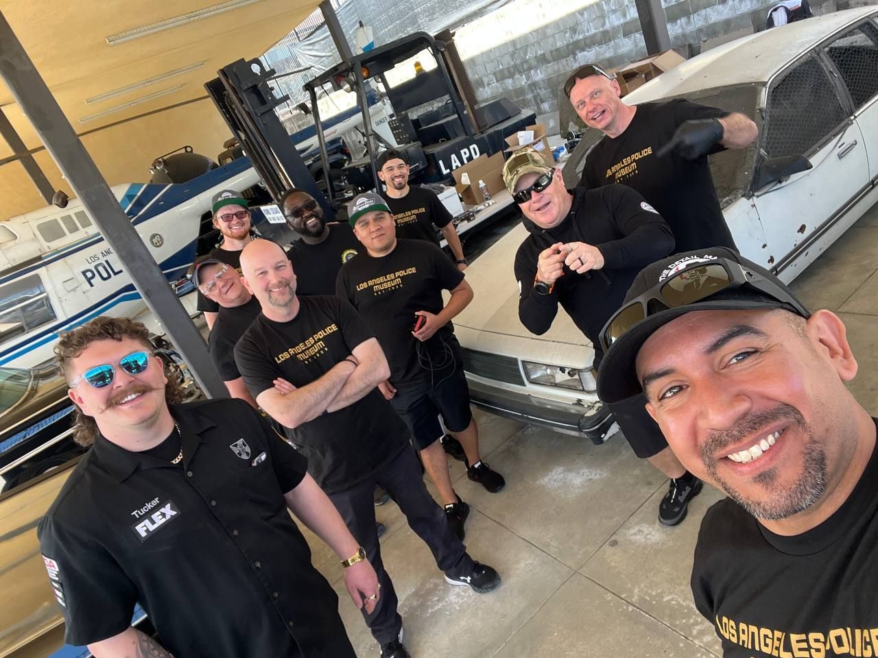 Group of mechanics smiling in a garage. Some wearing black shirts. Car & forklift in the background.