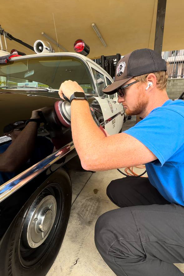 Man polishing a vintage black and white police car. He's kneeling and wearing sunglasses, and a blue shirt.