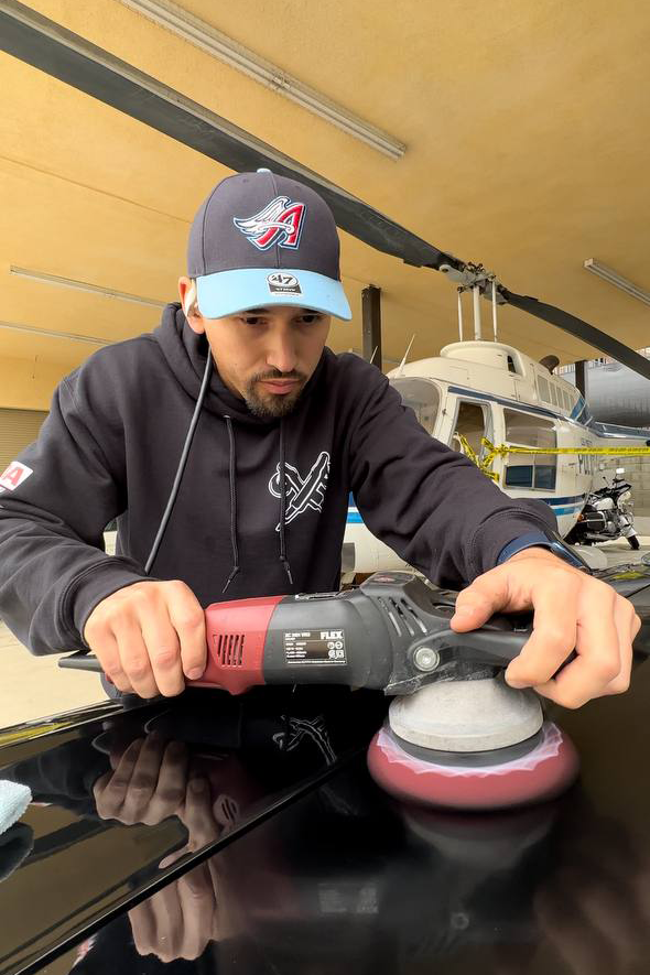 Man in a hat polishes a car with a machine, working in a hangar with a helicopter.