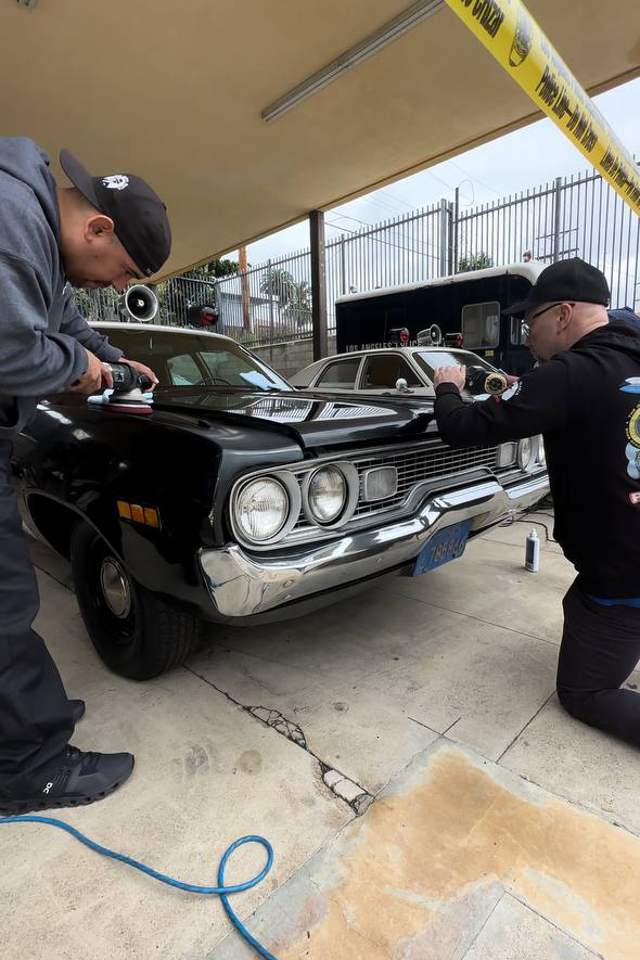 Two men polish a black vintage Plymouth police car, outdoors under a canopy.