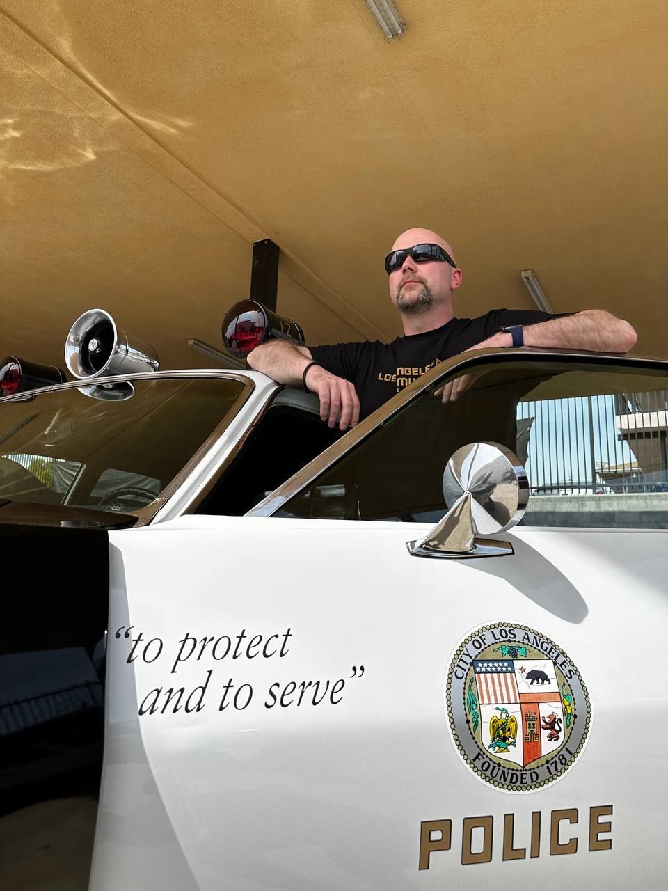 Man in sunglasses leans on a vintage LAPD police car, 