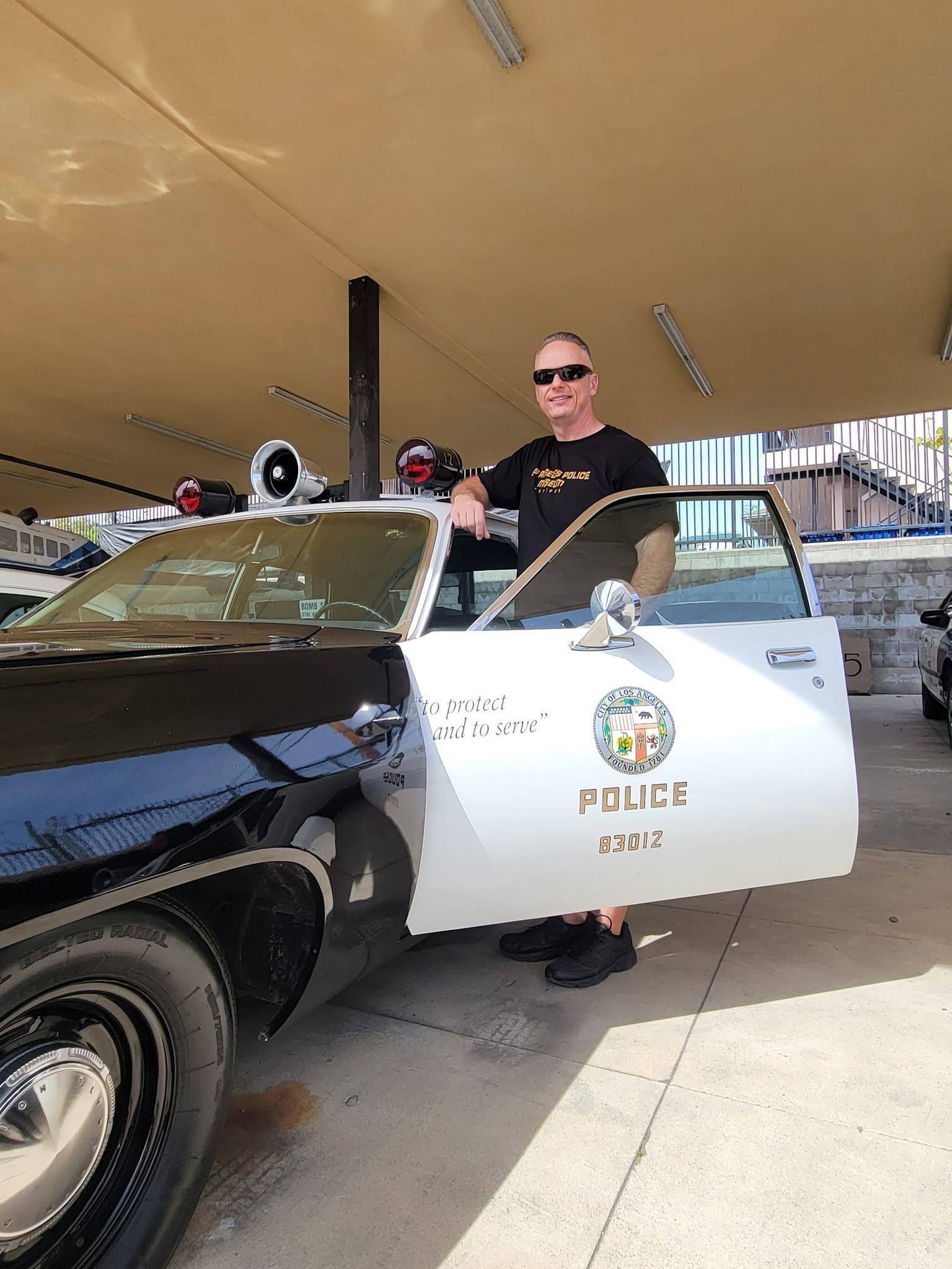 Man standing next to a black and white police car, under a canopy, with door open.