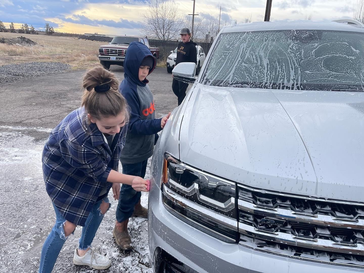 Three people washing a silver SUV with suds outside. Overcast sky, rural setting.