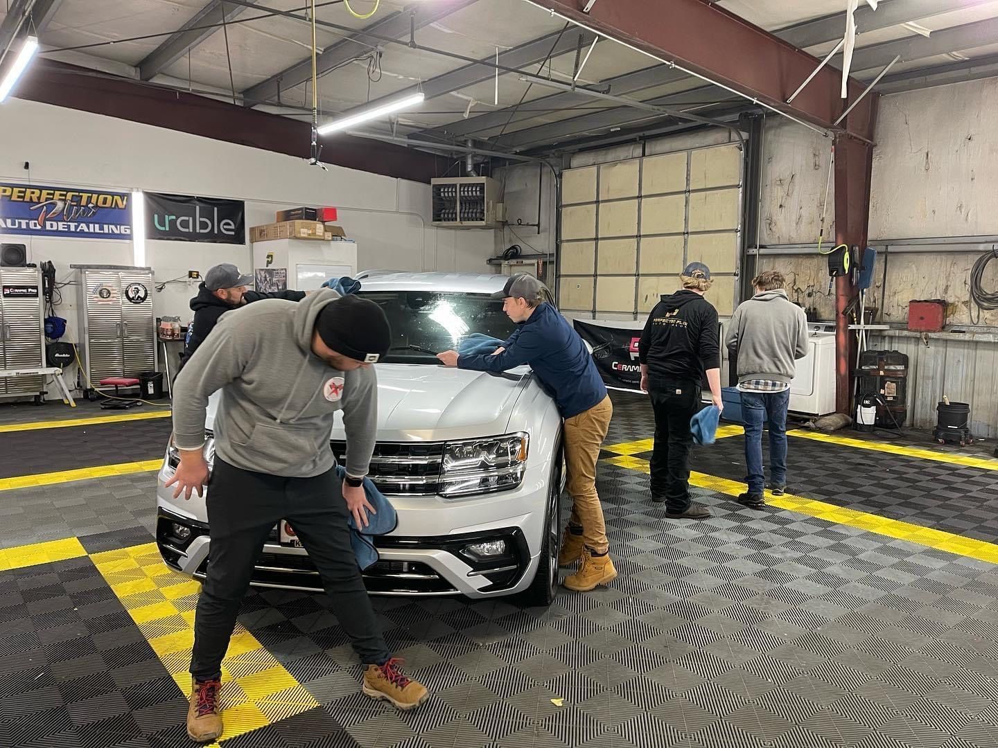 Five people cleaning a white SUV inside a garage with black and gray tile flooring.