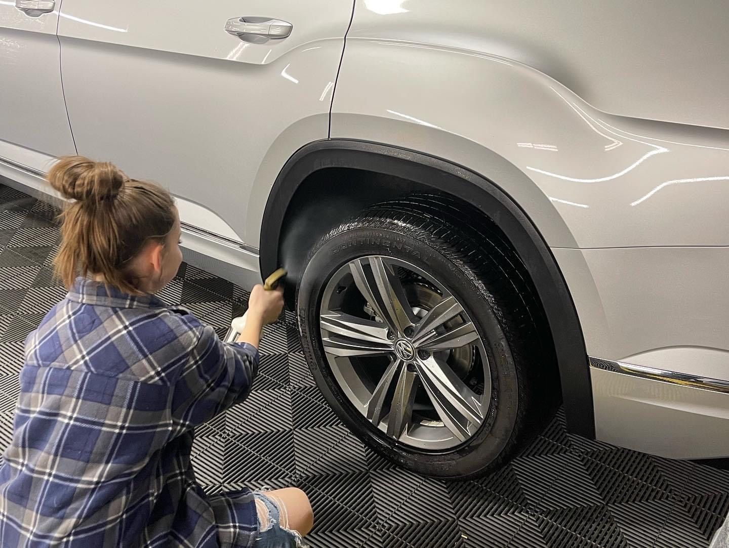 Woman washing a car wheel with a brush in a garage; white car, plaid shirt, black trim.