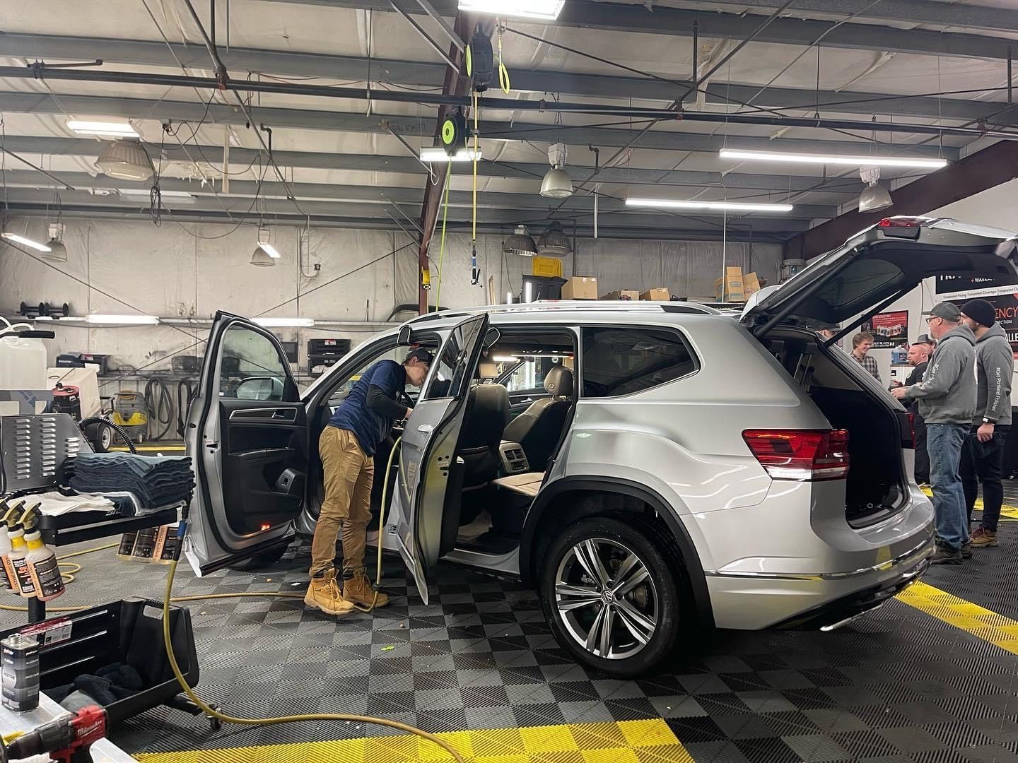 People working on a silver SUV in an auto shop with open doors and hatch.