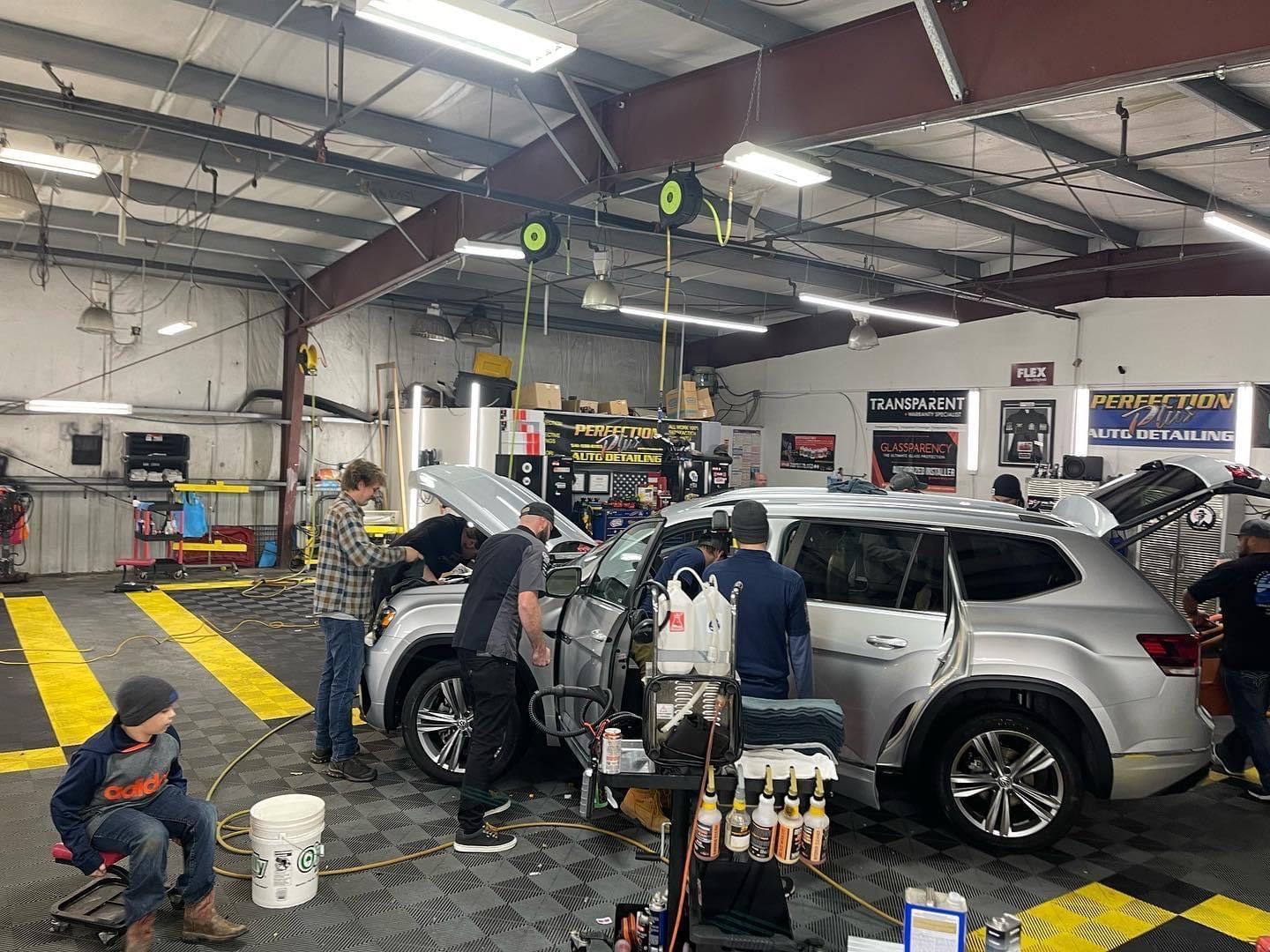 People working on a silver SUV inside a car detailing shop, with tools and equipment around.