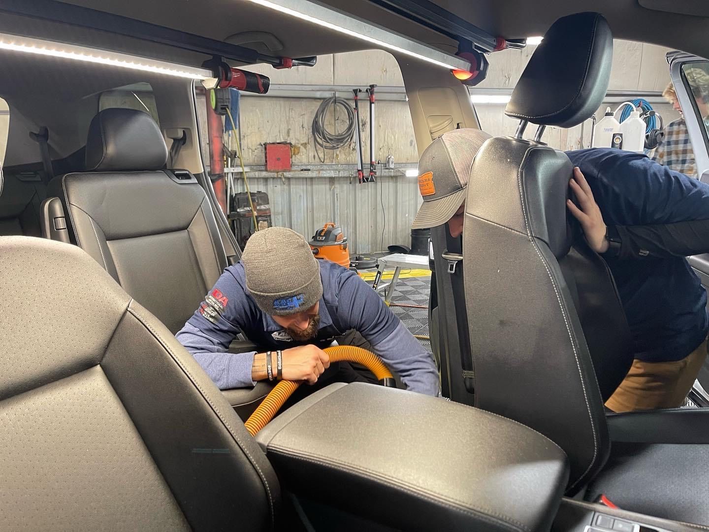 Two men working inside a car, likely repairing or cleaning. Garage setting with tools visible.