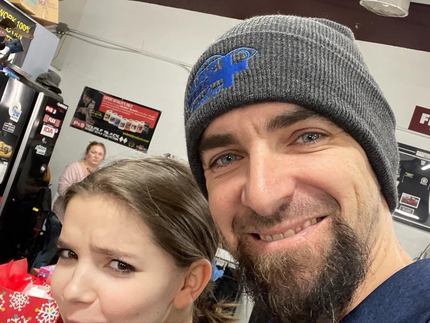 Man in beanie and girl make faces in front of vending machines, woman in background.