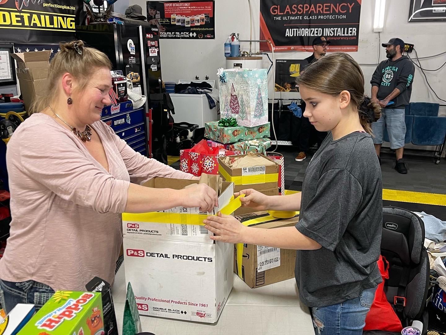 Woman and girl packing boxes with gifts in a garage, other people and wrapped presents visible.