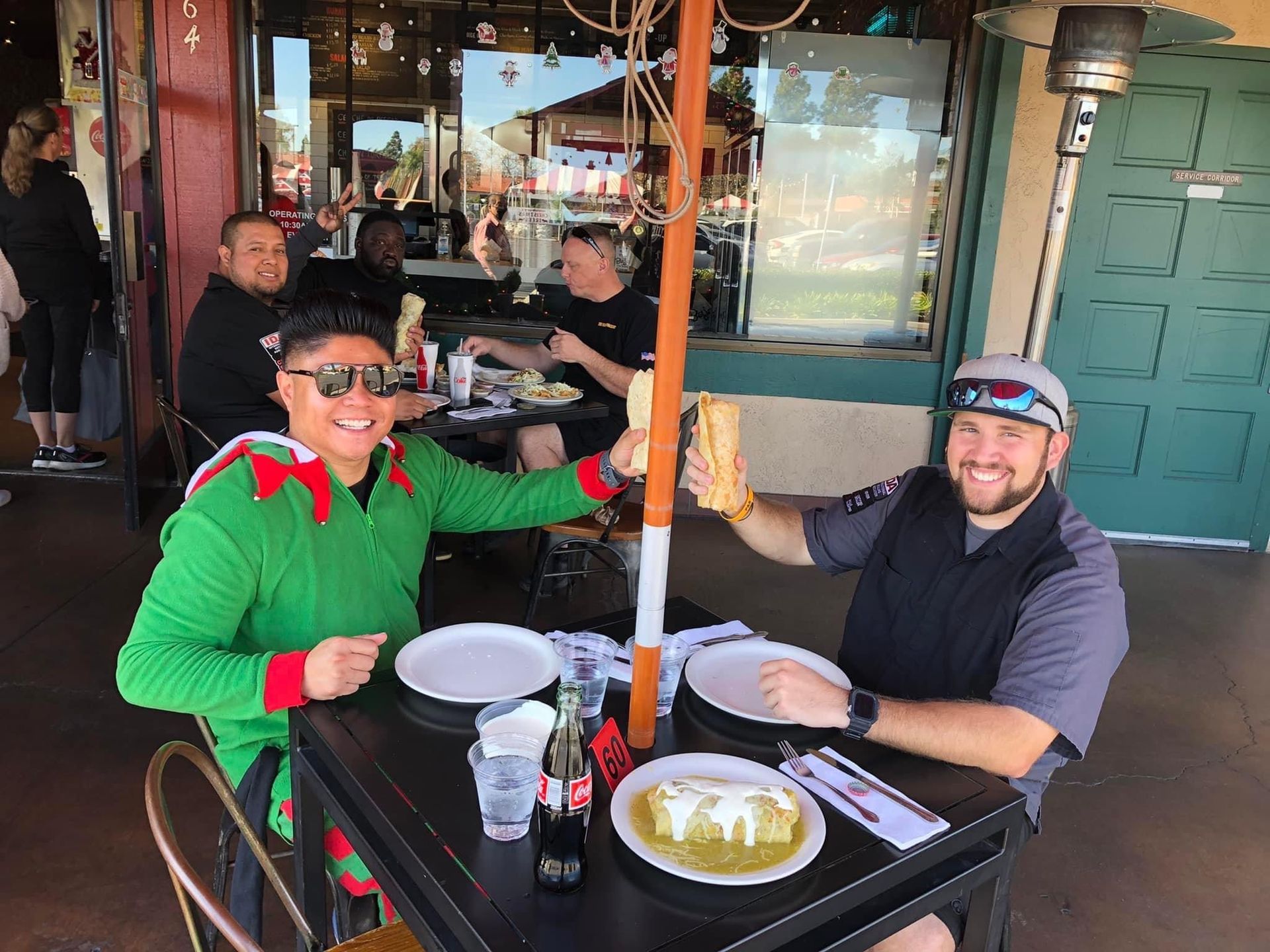 Two men in green and grey, toasting sandwiches outside a restaurant. Others eat in the background.