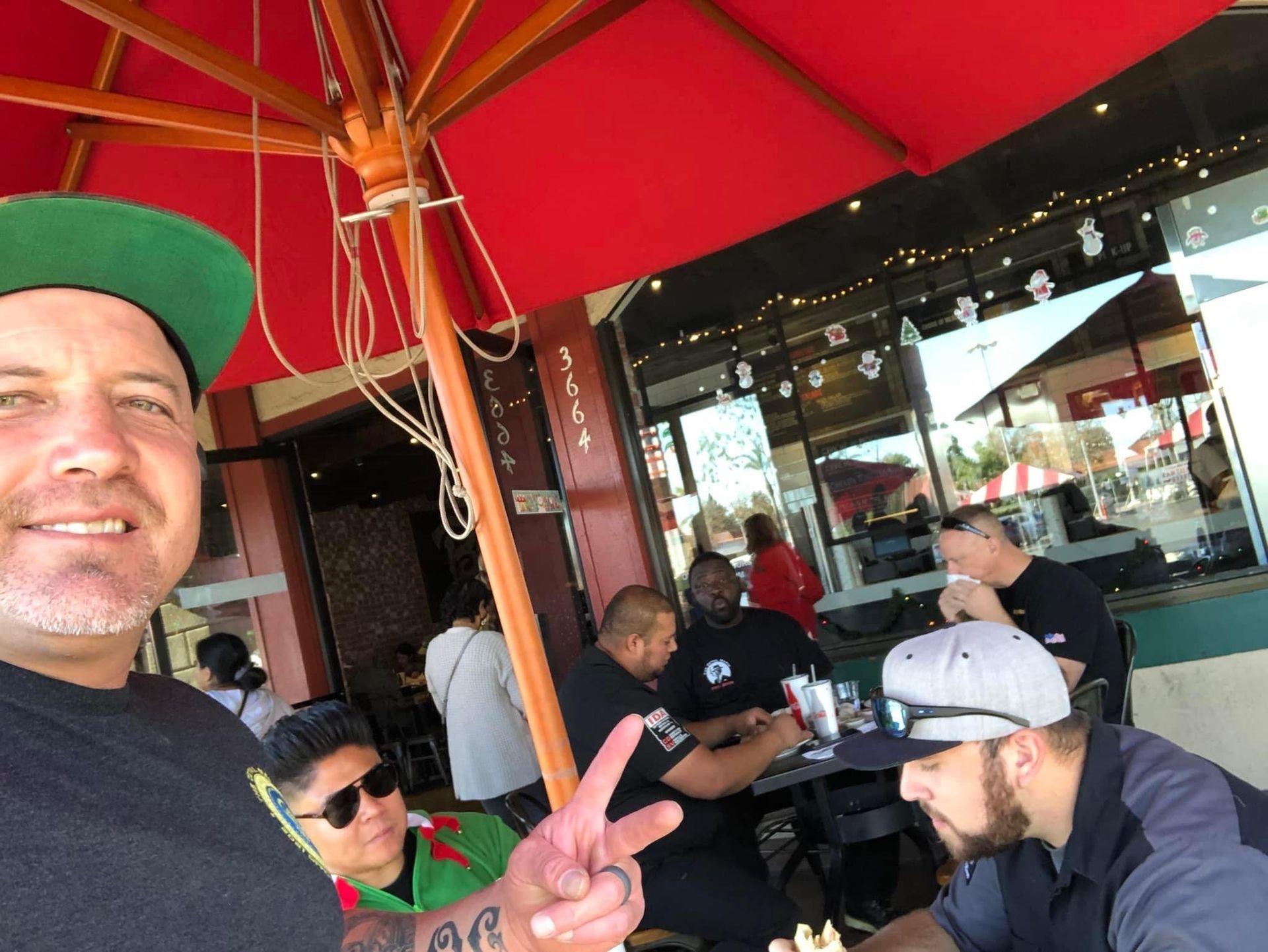 Man taking a selfie with group at outdoor cafe; red umbrella; people seated at tables; sunny day.