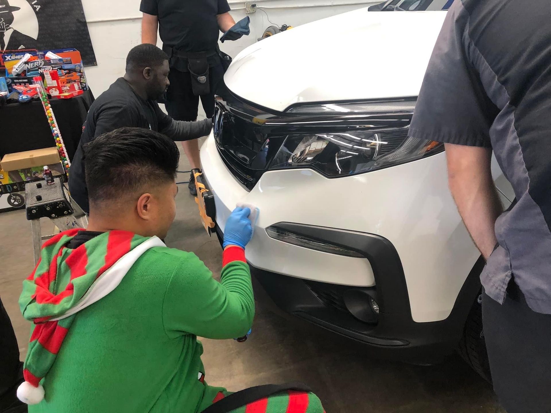 People applying film to the front bumper of a white car in a garage. One person wears an elf costume.