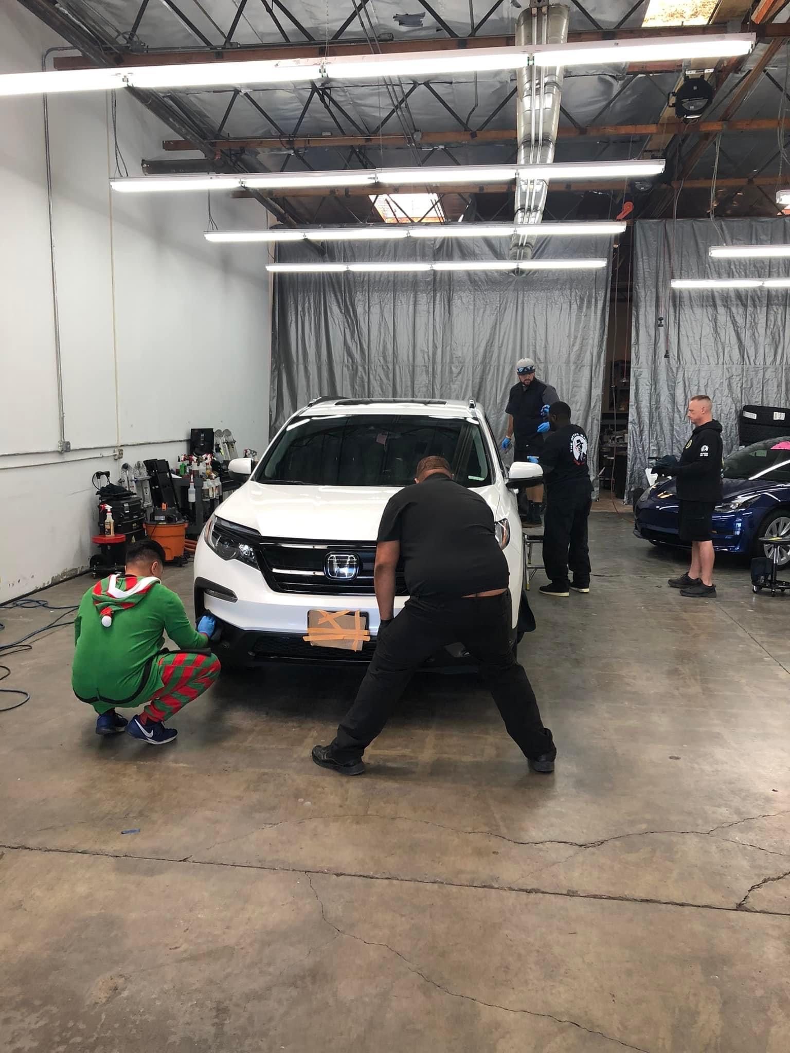 People working on a white car in a garage, one is wearing a Christmas sweater.