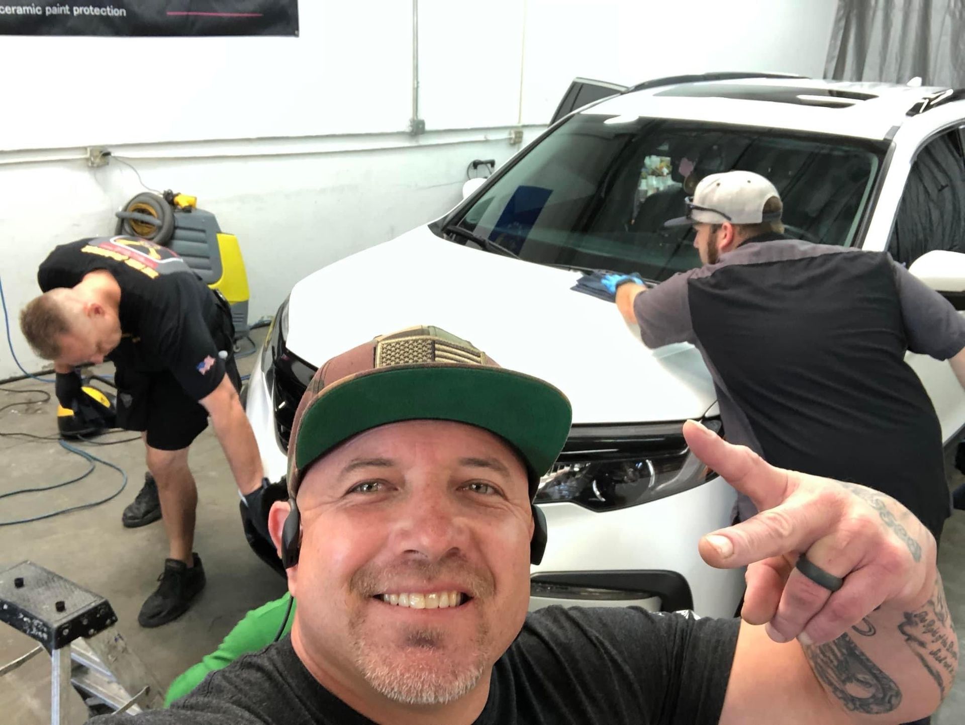 Man smiling, taking selfie, two others working on white car in garage.