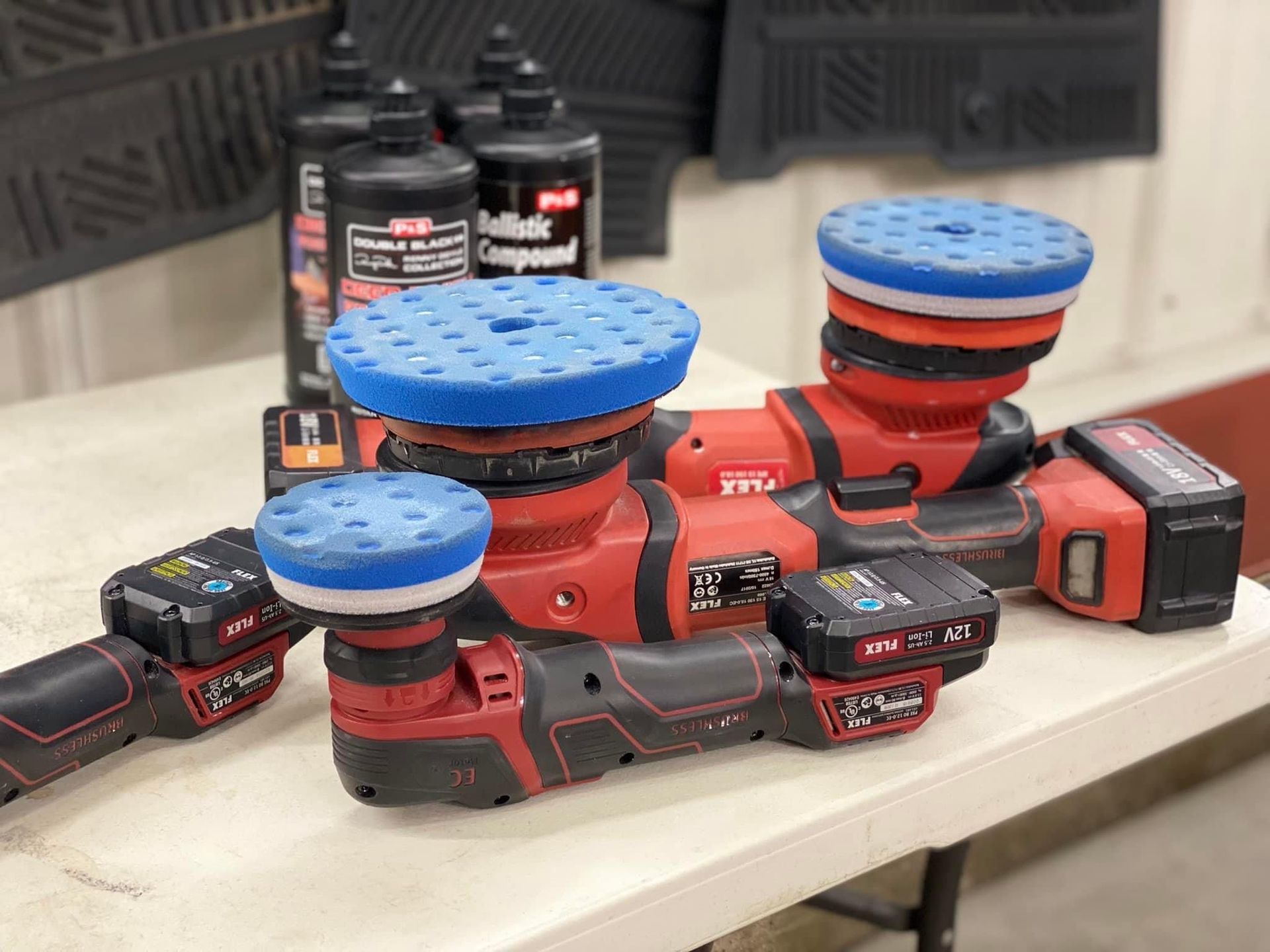 Three red and black polishers with blue and red pads on a table. Bottles of liquid in the background.
