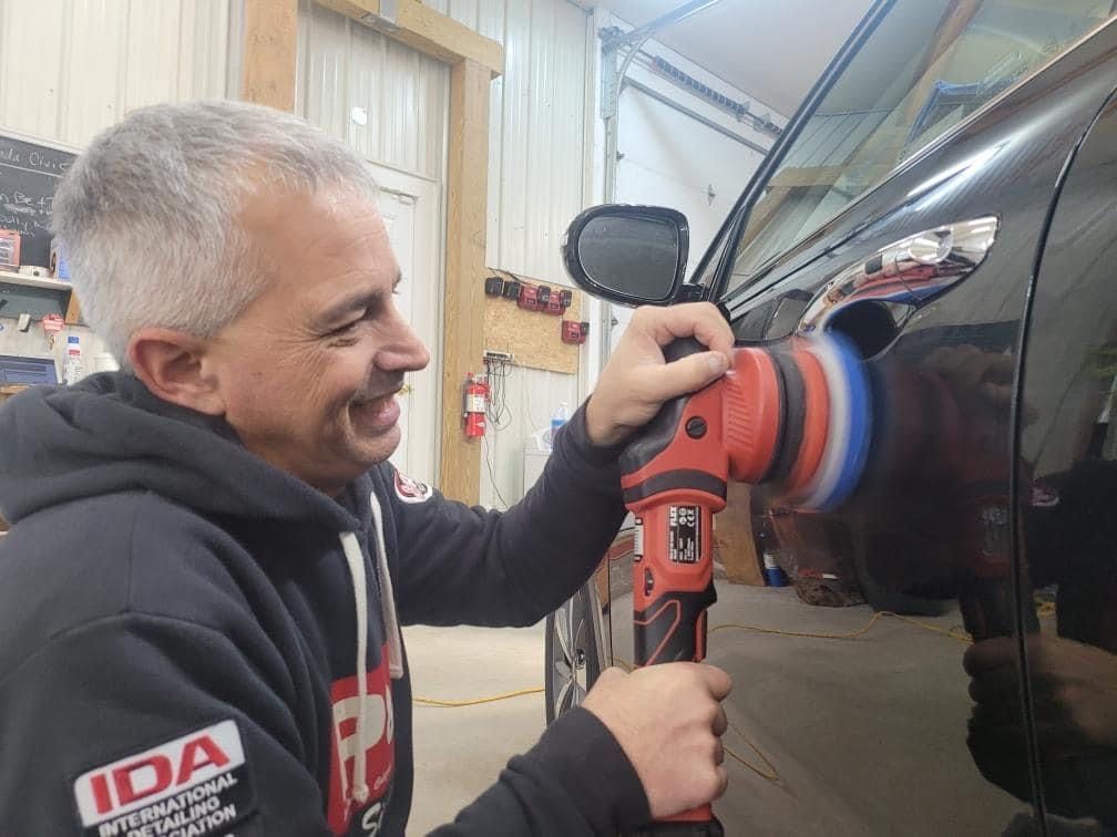 Man polishing a black car door, smiling. He's in a garage setting, holding a red and black polisher.