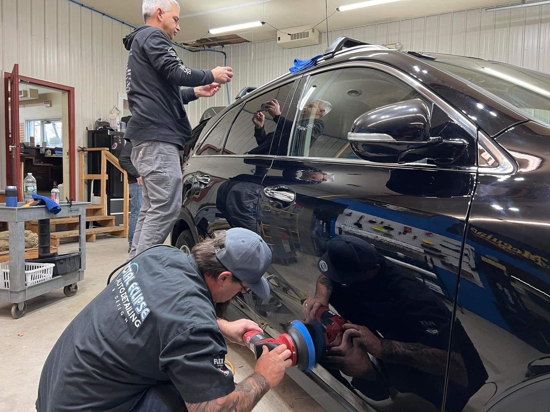 Two men detail a black SUV inside a garage. One polishes the side with a machine while the other takes a photo.