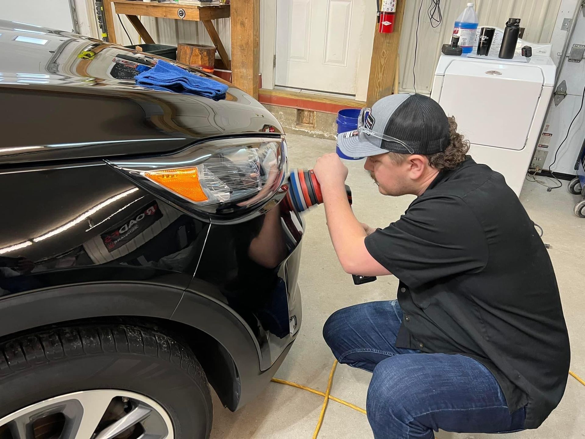 Man polishing car headlight outdoors, wearing cap, black shirt, and jeans.