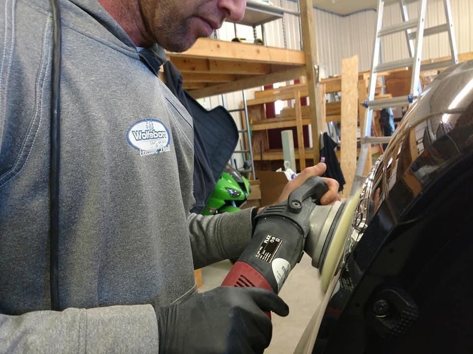 Man polishing a black car with a power buffer in a garage, wearing gloves and a gray sweatshirt.
