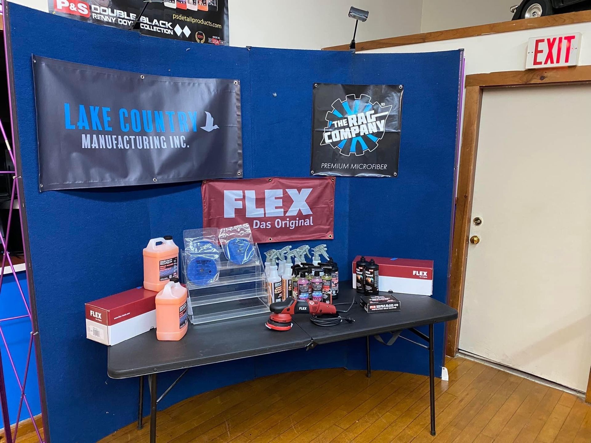 Display of car detailing products and banners at an event, featuring blue backdrop and black table.